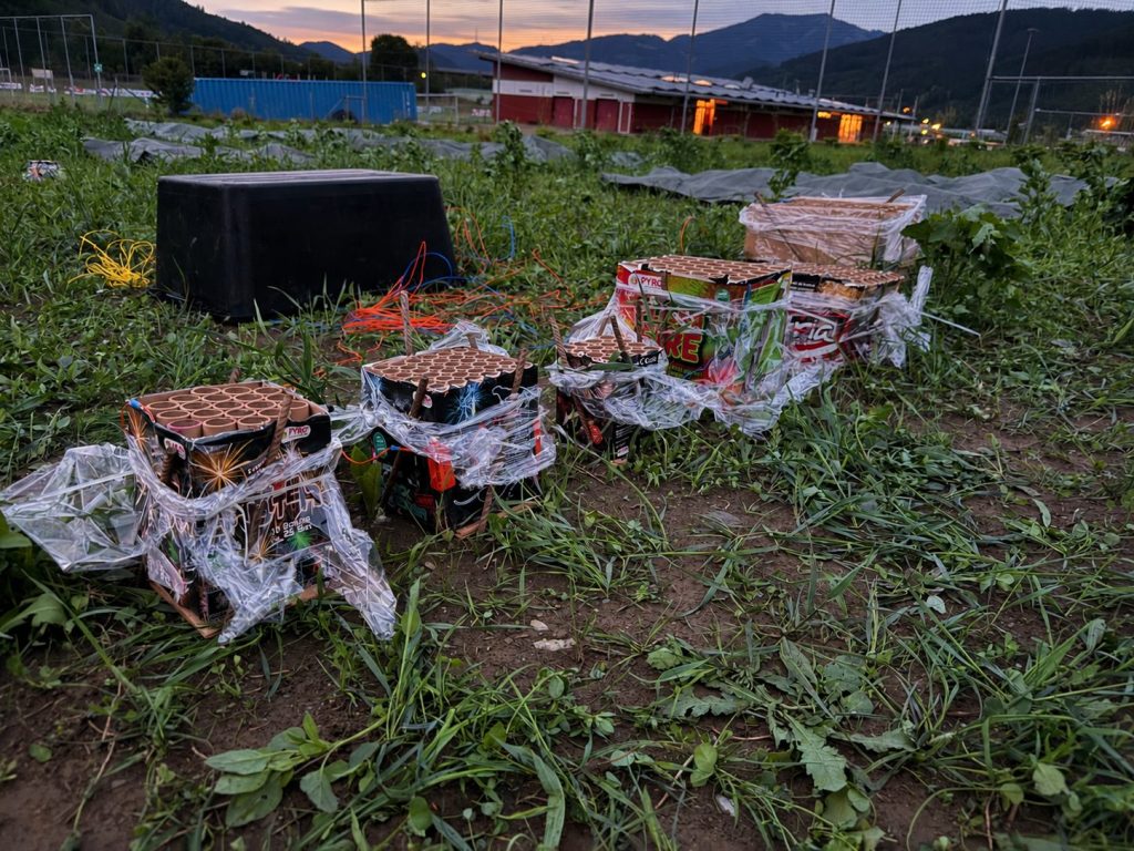 Alternativtext: Mehrere vorbereitete und verkabelte Feuerwerksbatterien auf einer Wiese bei Abenddämmerung.