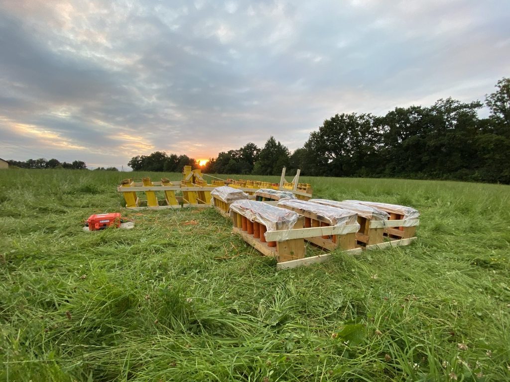Vorbereitetes Feuerwerk mit Abschussgestellen und Zündanlage auf einer Wiese bei Sonnenuntergang.