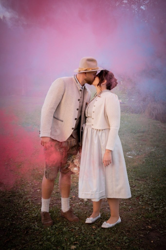 Küssendes Brautpaar umgeben von rosa und violetten Raucheffekten bei einer Hochzeit.