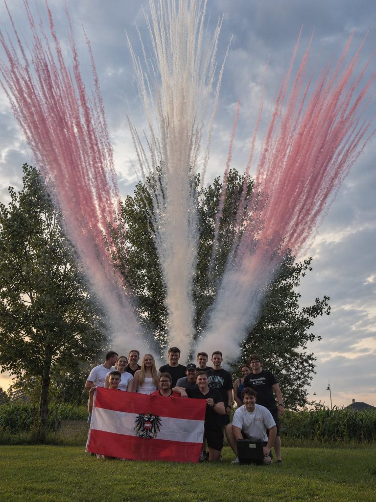 Gruppe vor einem rot-weißen Tagesfeuerwerk mit Raucheffekten im Freien.