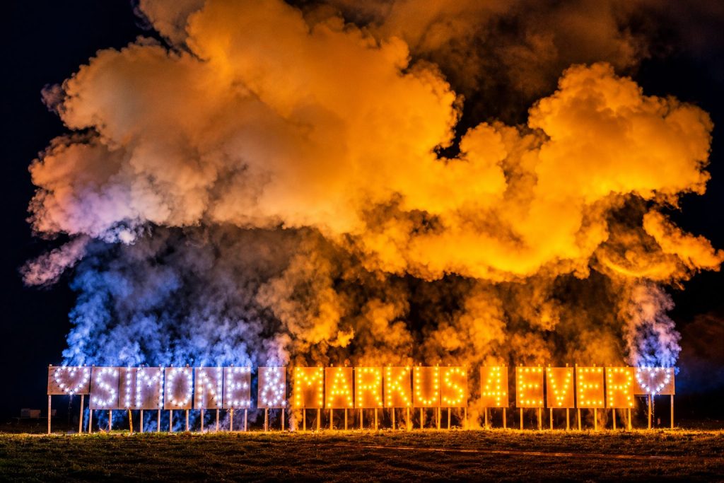 Lichterbild mit den Namen Simone und Markus sowie ‚4 EVER‘, umgeben von farbigen Raucheffekten bei Nacht.