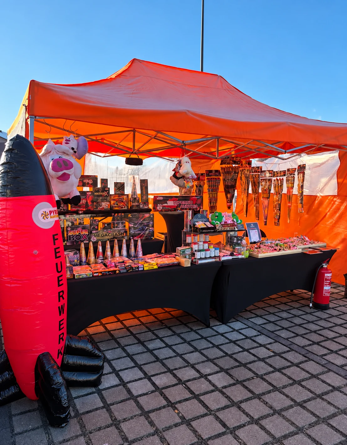 XP-Fire Verkaufsstand unter orangefarbener Plane mit Feuerwerk, Glücksbringern und großer Dekorakete.